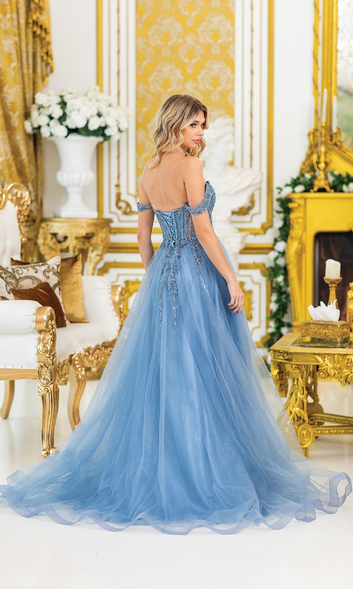 A woman in the Lace-Up Long A-Line Prom Dress: Dancing Queen 4490, featuring a light blue off-the-shoulder corset-style bodice, stands in an ornate gold-accented room, facing away from the camera.